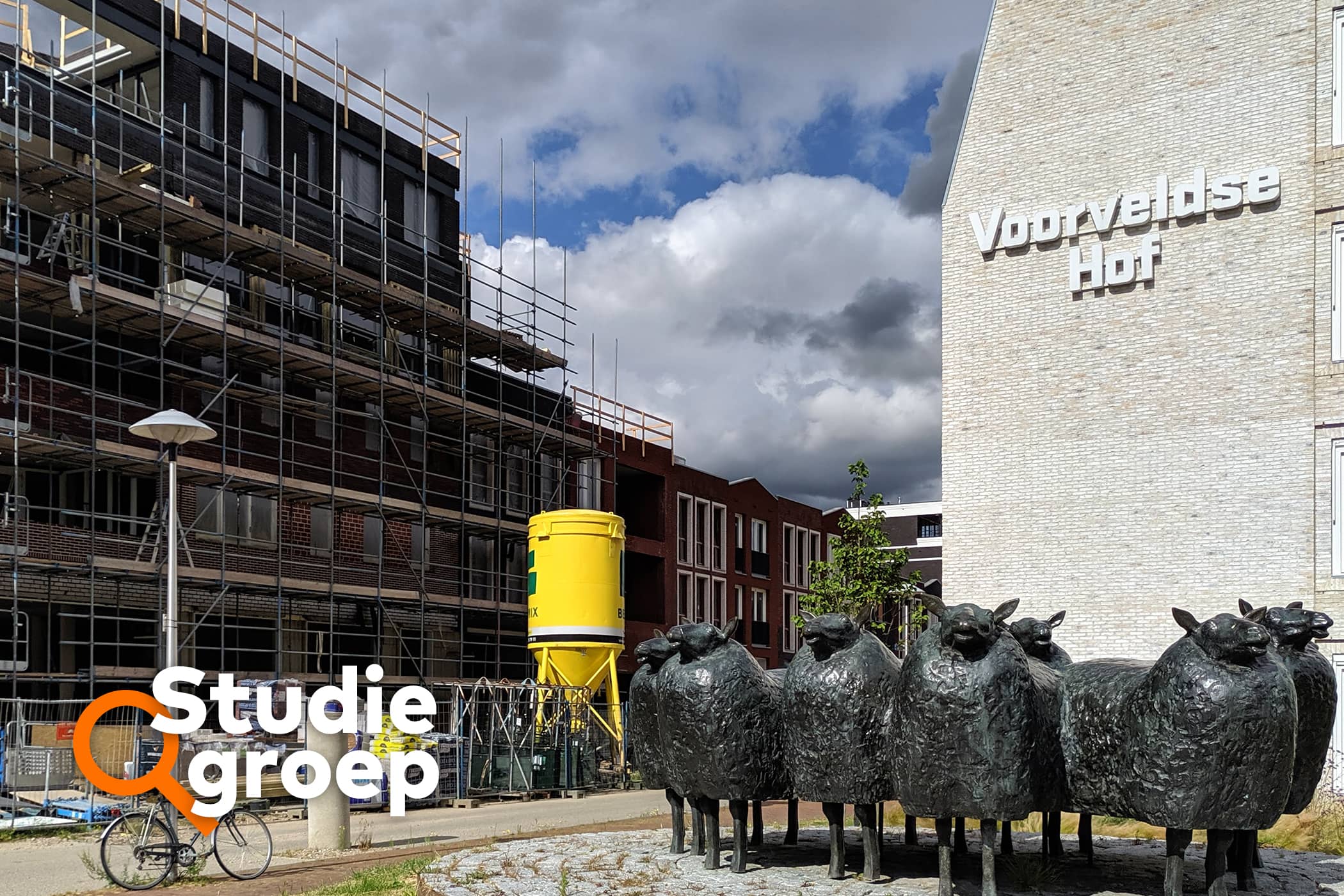 Een plein op straat met beelden van schapen. Erachter staat een wooncomplex in de steigers. Rechts staat een gebouw met erop de woorden 'Voorveldse hof'
