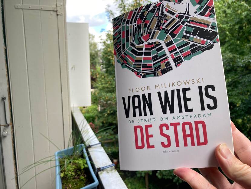 een hand houdt het boek 'Van wie is de stad' vast. Op de achtergrond is een balkon te zien