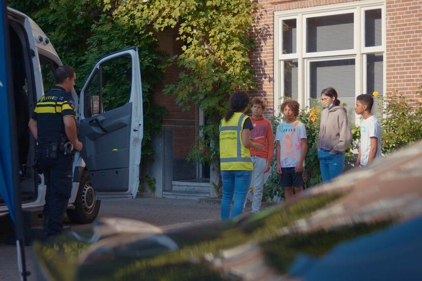 Vier jongeren staan naast de bus, vrouw praat tegen hen. Politieman staat naast de bus